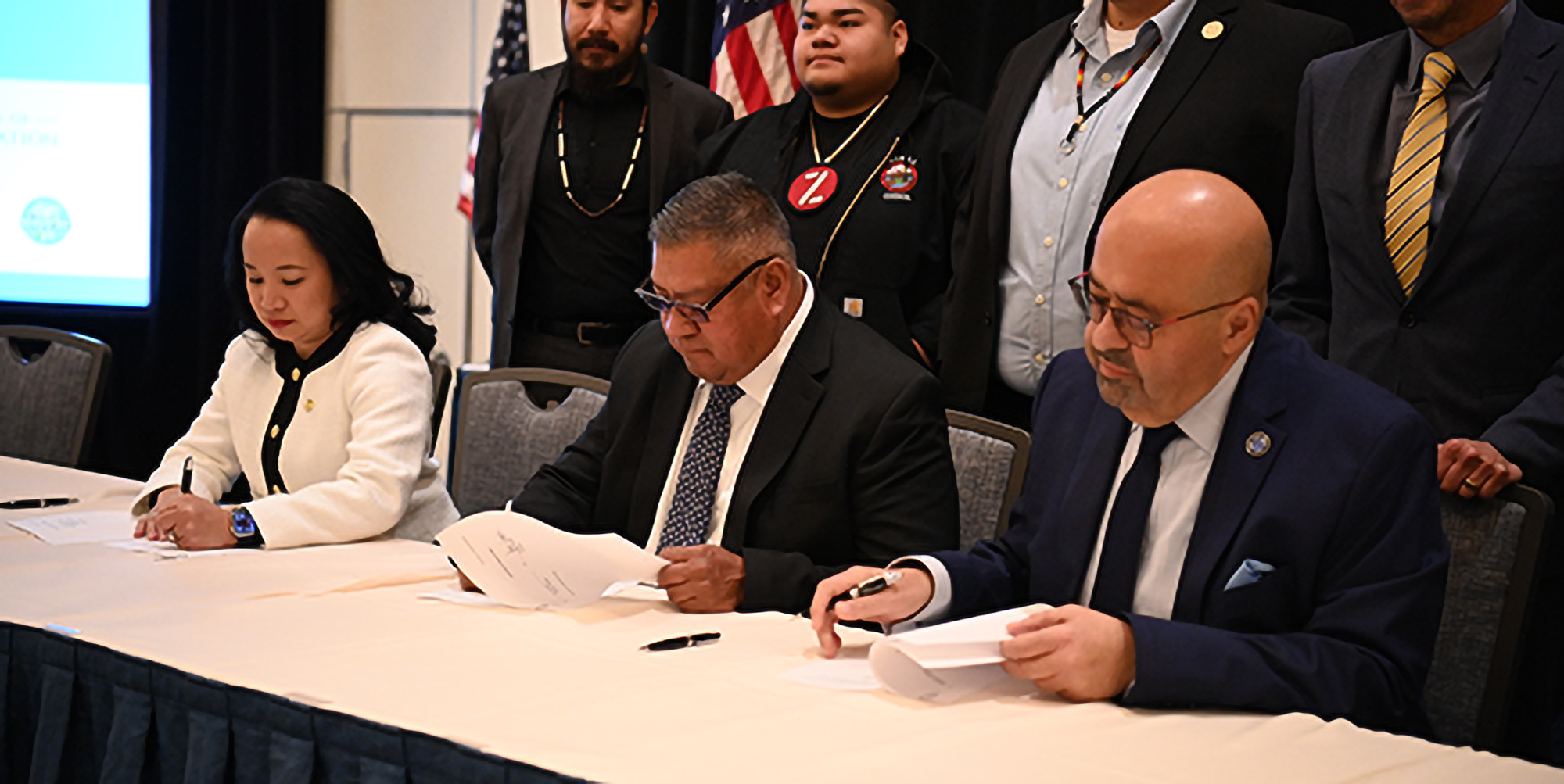 Three people signing agreements with people standing behind them.