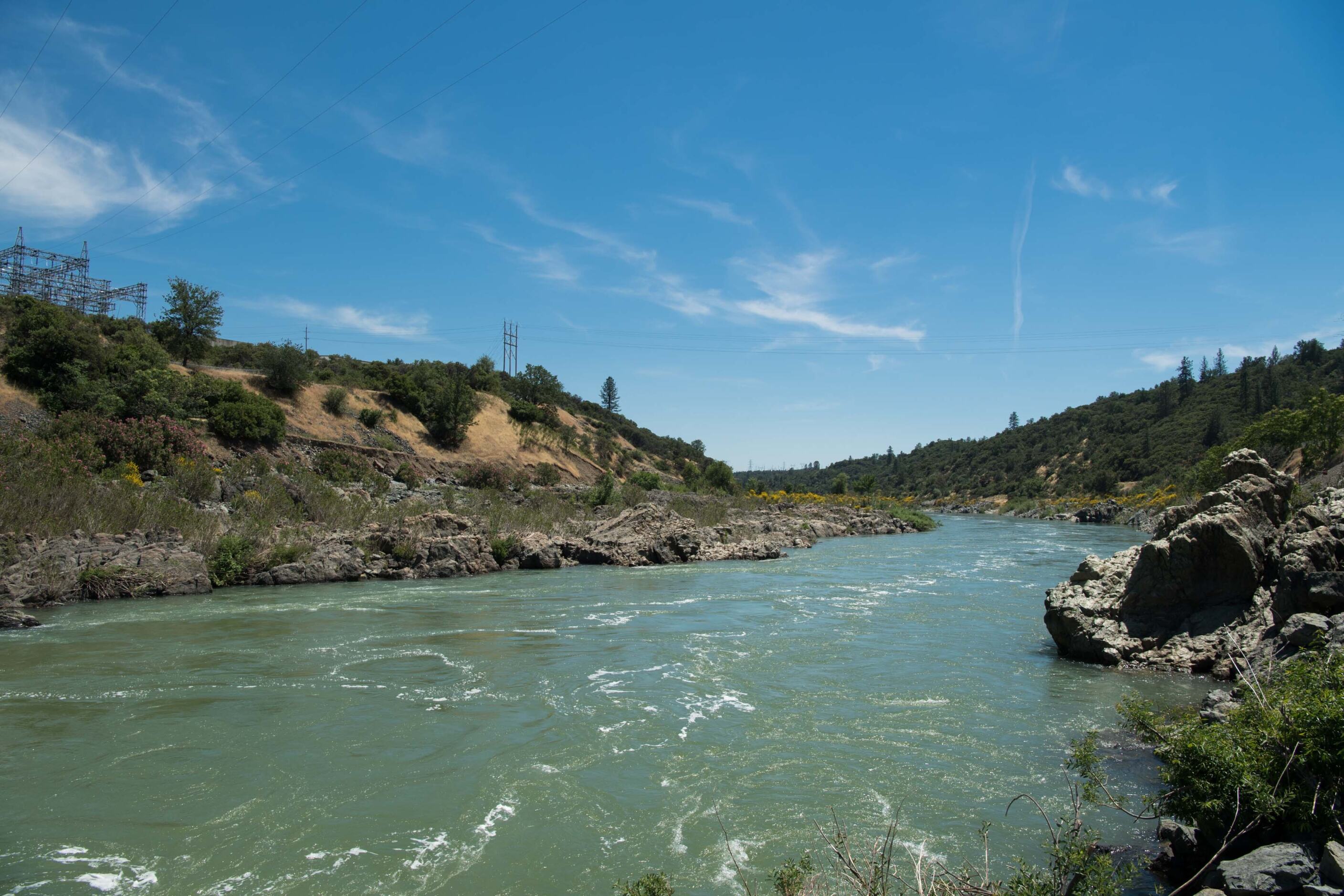 The Sacramento River running through brush and trees with high-power line towers on the hill.