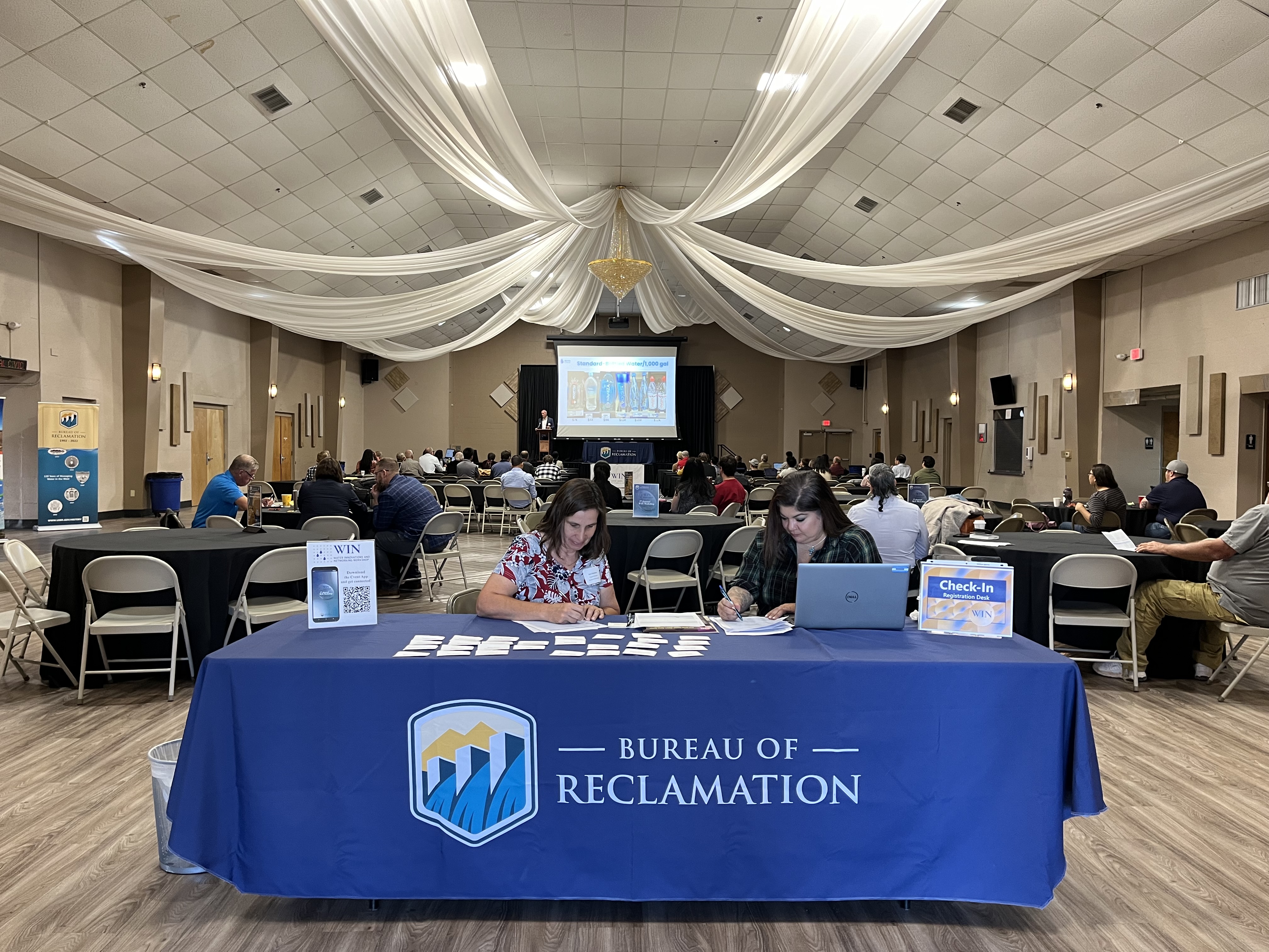 Two AAO staff seated at registration table fitted with blue Reclamation tablecloth. In background the Civic Center decorations and attendees visible.