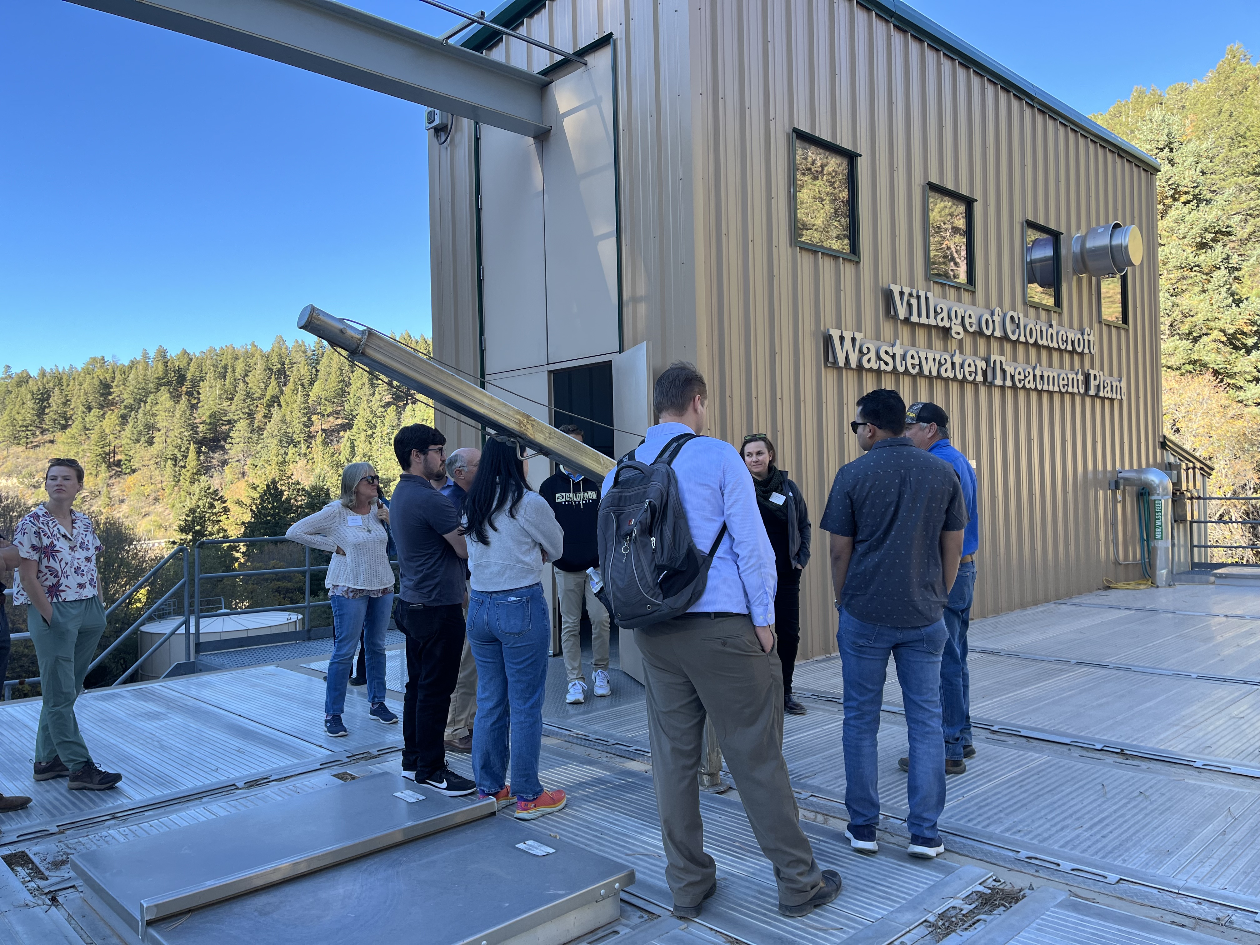 Several people gathered at the Village of Cloudcroft Wastewater Treatment Plant. The membrane treatment facility is in the background and is a brown metal building. Also visible are evergreen trees on the surrounding mountains.