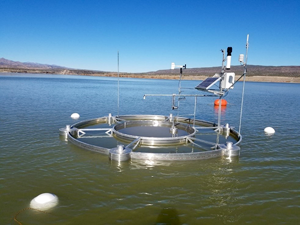 Daytime photograph of a floating evaporation pan partially immersed in a body of water surrounded by land. The unit displays various panels and antennae for gathering and storing reservoir data.