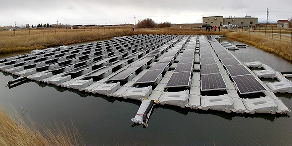 Daytime photograph of floating photovoltaic solar panels partially immersed in a body of water surrounded by land.