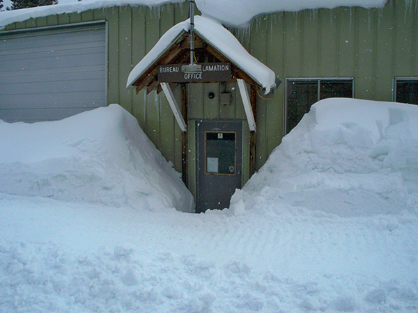 A door to a tan building is shown with snow piled higher than the door on either side.