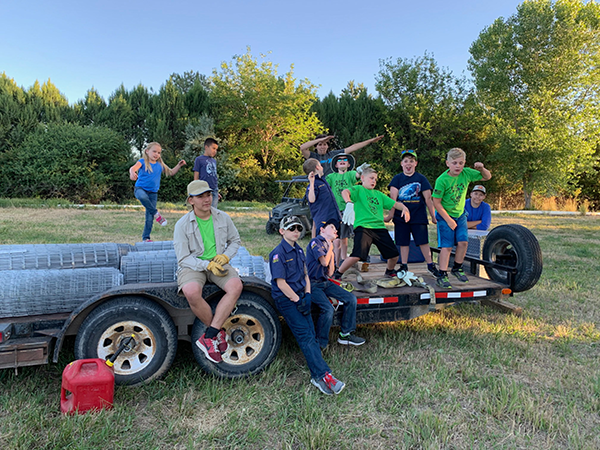 A group of 11 boy scouts and cub scouts pose with  tree caging equipment, while making funny faces and poses in a field at Grand Junction Wildlife Area around sunset.