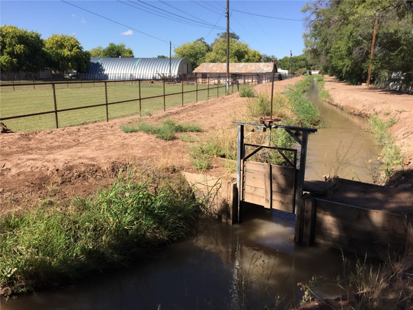 View of a canal check structure and the canal running next to a fenced field and a building in the background.