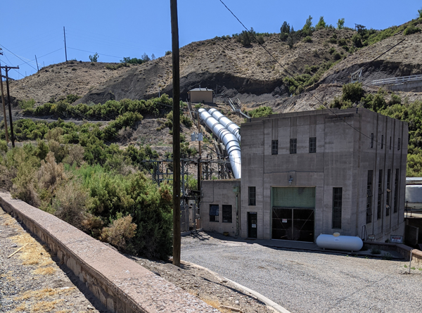 View of the Grand Valley Powerplant with two silver penstocks running up the hill behind the powerplant building. 