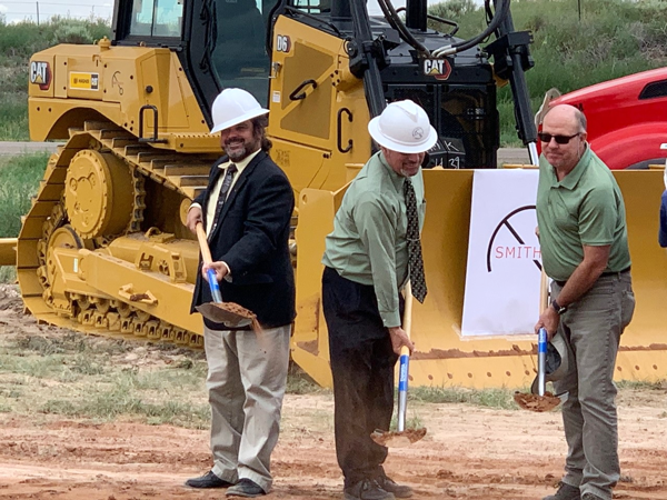 Closeup view of three professionals in hardhats turning dirt with shovels. Several pieces of heavy equipment are parked in the background. 