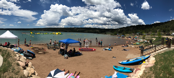 Brown sandy beach with paddle board and kayaks, two tents and people sitting on the sand. Out on the lake you can see a yellow and blue inflatable play area and it is a sunny day with blue sky and white clouds.
