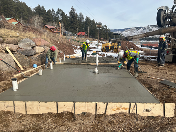 Four contractors in hard hats work on concrete forming of a rectangular concrete pad with a concrete truck next to the pad on a clear winter day