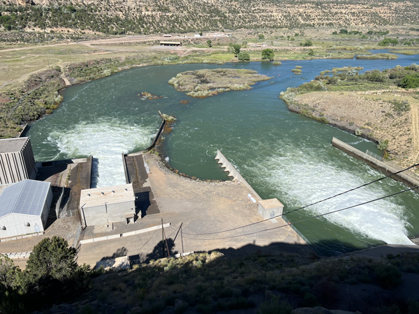 View from the top of Navajo Dam looking downstream into the San Juan River. There are three buildings on the left side of the photo and the river is blue with mountains in the background