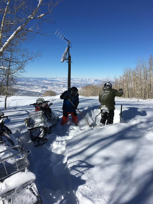 Two men in snowmobile gear and helmets use shovels to scoop snow out of the pipeline structure. Snow is all around then and two snow mobiles are parked on the left side. It is a sunny day with blue sky.