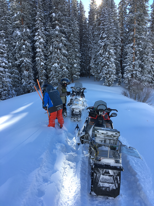 Two men in snow mobile gear and helmets with measuring gear surrounded by snow and pine trees and their snow mobiles near by on top of the small dam.