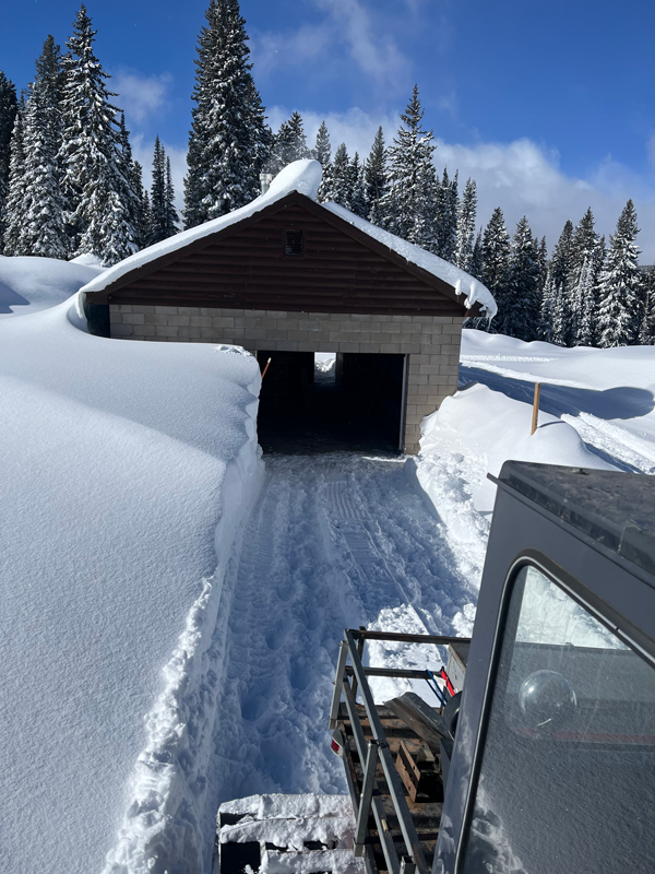 A brown and brick building with a snowplowed path leading to the open garage door, part of the snow cat is visible as it makes its way down the path with several feet of snow on each side.  It is a sunny day with blue sky and snow-covered pine trees in the back.