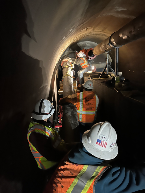 Four constructions workers lift a 12-inch steel piece of pipe into position inside of the outlet works tunnel.