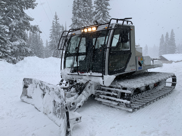 – a gray snow cat with a plow attached to the front and lights on.  It is lightly snowing, and the snow cat is moving down the path of snow.