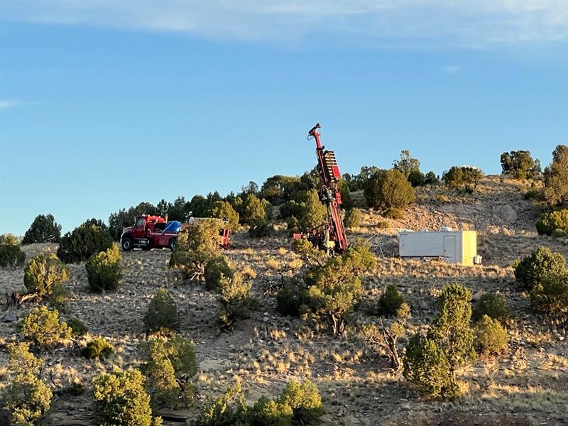 On the hillside there is a large read truck and red drill rig next to a small white maintenance building.  It is just after sunrise with blue sky in the background