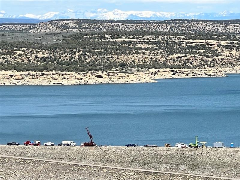 Far off photo of Navajo Dam with Navajo Reservoir and rocky landscape and shrubs, with snowcapped mountains in the distance.  A long line of work vehicles, construction equipment, and drilling equipment are visible along the dam road.