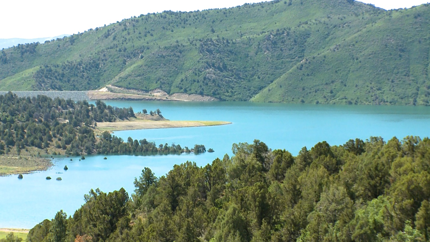 Blue water of Lake Nighthorse with a green mountain top in the background and green trees and shrubs surrounding the shoreline